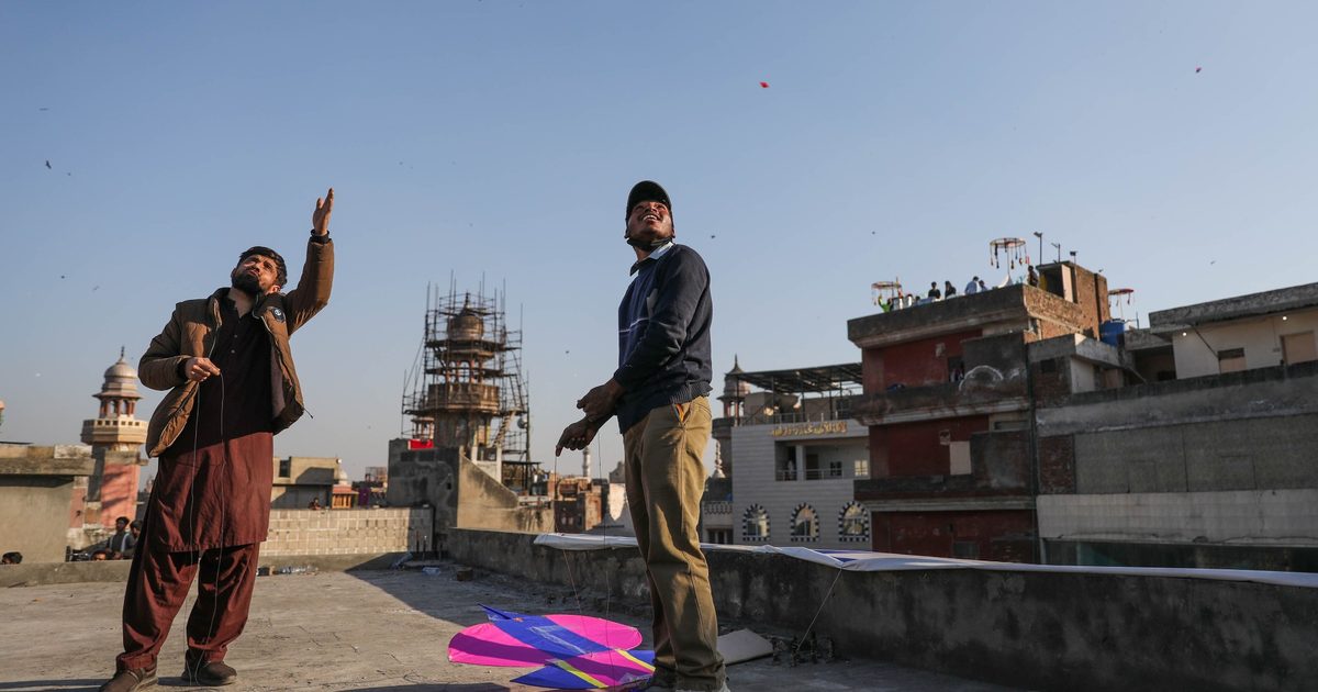 Two men flying kites from a Lahore rooftop with a low parapet, the old city behind them
