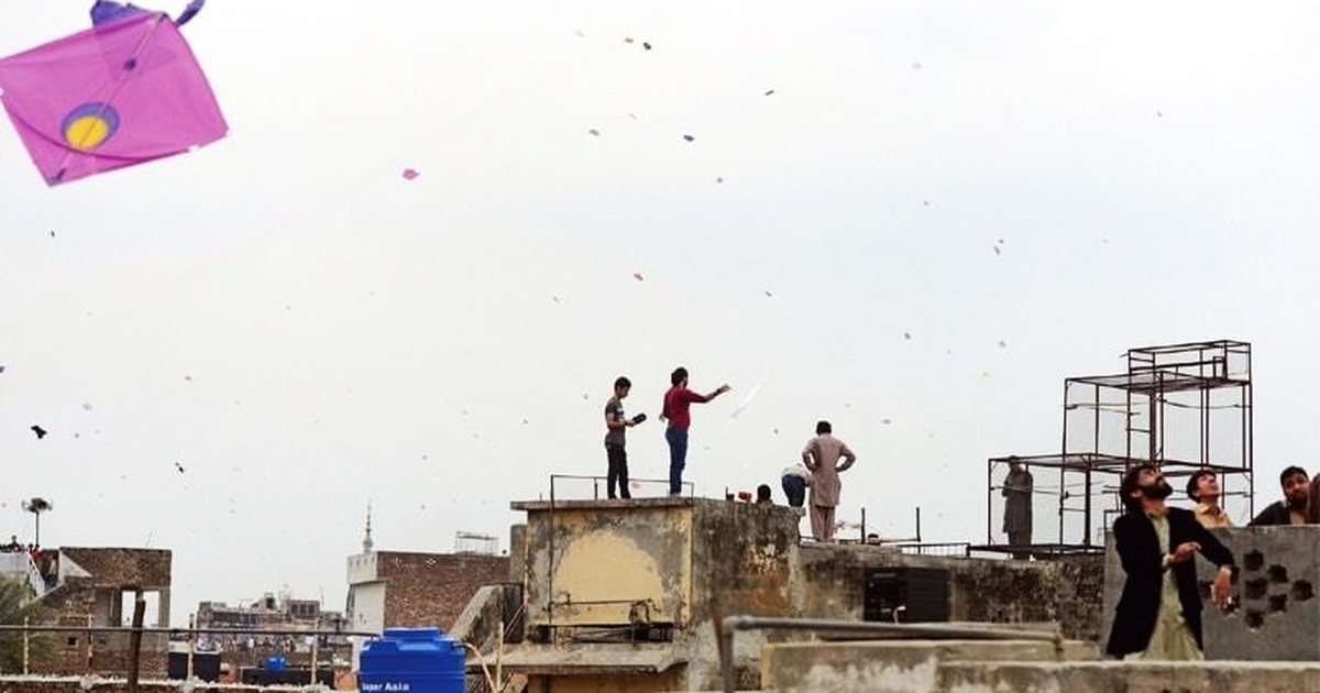 Young flyers on Lahore rooftops with kites overhead, some of the roof edges unwalled